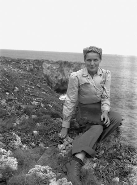 Leota Kelly, director of the Red Cross's Pacific Athletic Club, posing for a picture by the shore in Tinian at the site of the new Red Cross Club. Kelly is wearing cuffed pants, a button-up blouse and shoes, with her hair up in a scarf. She is holding a large black folder.