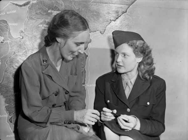 Two nurses, Leona Jackson, left, and Mary Rose Harrington Nelson, right, return to Guam after being imprisoned in Los Banos, Philippines, for over two years. Jackson is wearing a dress with large buttons. Nelson is wearing a cap and dress suit uniform, and holding a long pen-like object. Behind them is a map of Guam painted on the wall. Leona Jackson went on to become the third director of the United States Navy Nurse Corps in 1954.