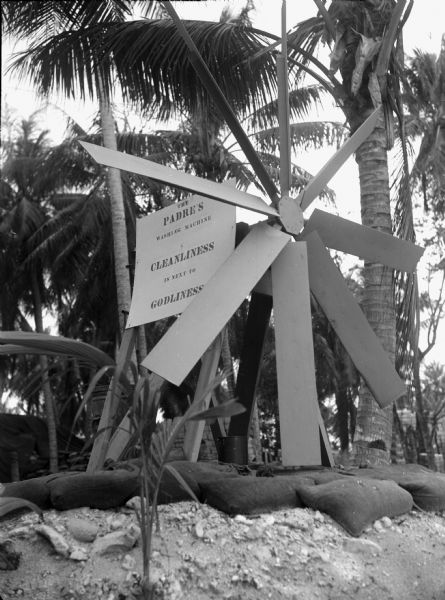 A washing machine run by a windmill in Ulithi. Behind the large propellers of the machine is a sign that says: "The Padre's washing machine---Cleanliness is next to Godliness". Behind that are palm trees. Under the windmill are sand bags and a trash can.