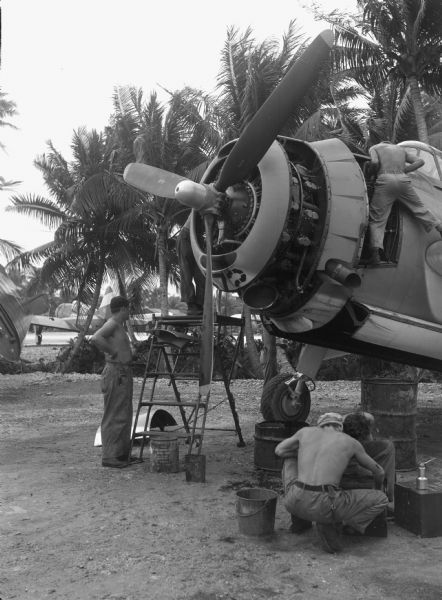 Men working on a Marine Aircraft Group plane at Ulithi airport. One man is standing near the nose of the plan, looking inside. He is shirtless and wearing light-colored pants. Two shirtless men are squatting on the ground below. The third man, also shirtless, is standing shirtless by a platform ladder that another man has climbed up. Buckets and barrels litter the ground. In the background are palm trees and other planes.