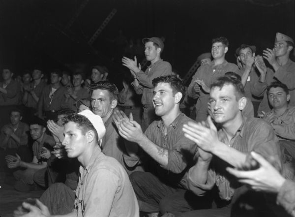 Surviving crew members of the USS <i>Franklin</i> watching an amateur vaudeville on their hanger deck, seven says after their disaster. The USS <i>Franklin</i> had maneuvered closer to the Japanese mainland than any other carrier and was bombed twice from above by a Japanese plane. There were 724 casualties. The remaining crew managed to save the ship. The men are clapping and many of them are smiling.