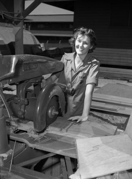 Private Winnie L. Cockrell, a carpenter from the United States Marine Corps Women's Reserve, at the Mauna Loa Ridge encampment in Honolulu. Cockrell is wearing a jumpsuit and smiling. In front of her is a large table saw and some partially cut wood.