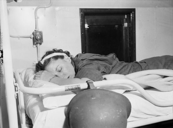 A nurse sleeping at battle stations aboard the USS <I>Relief</I> in the China Sea. The nurse is wearing a jumpsuit and has a piece of cloth tied in her hair. She is sleeping in a bunk. There is a helmet strapped to the side of the bed.