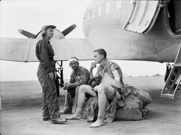 Flight nurse, Gwen Jensen, wearing a jumpsuit and a cap, standing and waiting with near an airplane with marine Private First Class George Berier, left, and marine Corporal John H. Kerr, right, from the 4th Division. The two men are both sitting on a pile of baggage while awaiting takeoff near Iwo Jima. Berier has a bandaged head. Kerr is wearing ripped clothing and has his arm in a sling.
