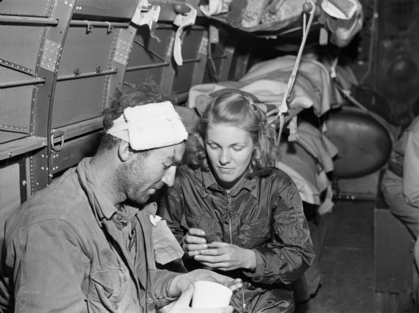 Flight nurse, Gwen Jensen, with Private first class George Berier. Berier's head is wrapped in gauze, and he is preparing to take medicine. Jensen is wearing a jumpsuit and watching Berier. They are inside an airplane and people in stretchers are behind them.