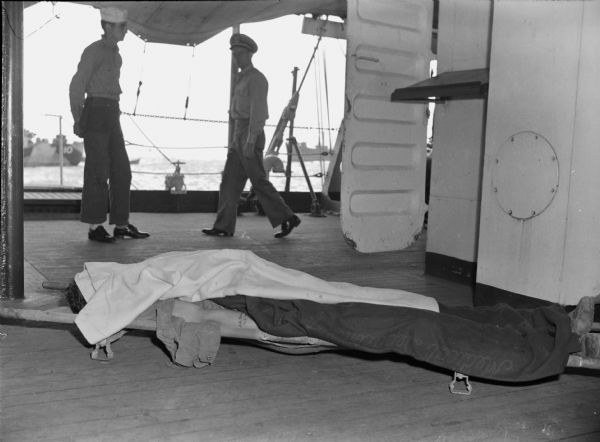 A body on a stretcher is lying on the deck of the USS "Samaritan." The body is covered by a blanket, except for an arm which has a small tattoo. Behind the stretcher a sailor is standing as a man in uniform is walking past. Other ships are in the background.
