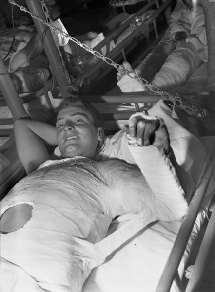 Private First Class George Pantaloni in a plaster cast aboard the USS <I>Samaritan</I>. He is lying in a cot and smiling. Behind him are more cots with other men on them. One man behind Pataloni is looking towards the camera.
