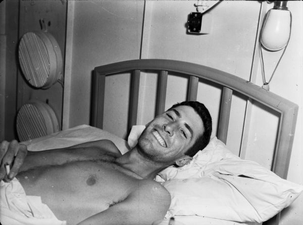 Second Lieutenant Louis Connick smiling for a photograph aboard USS <I>Samaritan</I>, a hospital ship. He is lying in a bed and holding a cigarette. He is shirtless and has a sheet pulled up to his waist.