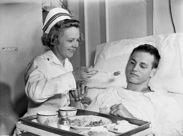 A nurse aboard the USS <I>Samaritan</I> feeding a wounded soldier. The man is sitting up in bed as the nurse is raising a fork full of food to his mouth. There is a tray of food in front of him.