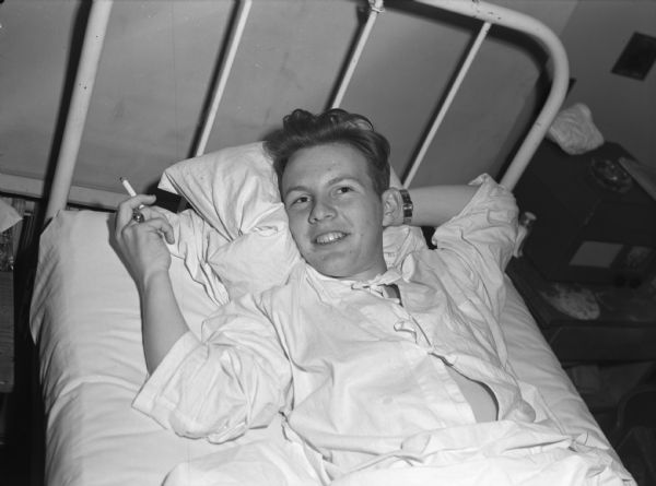 Private First Class George Bebbington, who was wounded in Iwo Jima, resting at the St. Albans Naval Hospital in the United States. He is smiling and relaxing in a bed with a cigarette in his hand.