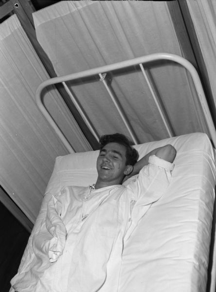 Private First Class William Fenton, wounded in Iwo Jima, laughing after being returned to the United States, to St. Albans Naval Hospital. He is lying down in bed and has one arm behind his head and one arm inside his shirt.