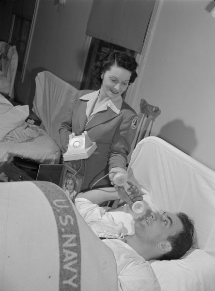 A woman with a badge marked "Bell Telephone System" is holding a telephone cradle while handing a wounded marine a telephone receiver. The marine is lying in bed with a U.S. Navy blanket, and beside him is a framed photograph of a woman.