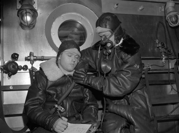 A flight nurse in an anoxic faint chamber, used to simulate high altitude flying with little oxygen, placing an oxygen mask over a male patient's face. He is writing on a clipboard but the writing has gone off the page. The man is wearing thick leather clothing and is almost closing his eyes. The nurse is also wearing thick leather clothing and already has an oxygen mask on. A woman is looking in the porthole window behind them. This photograph was taken at Alameda Naval Air Station in California.