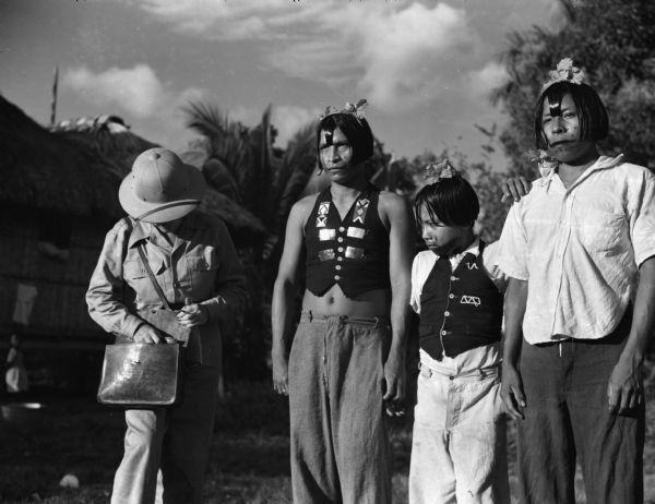 A Panamanian boy is standing between two indigenous Panamanian men. They are all wearing face paint and flowers in their hair. One of the men and the boy are wearing vests with pants. The other man is wearing a button-up shirt and pants. Beside them an American woman is searching in her bag. She is wearing a safari hat and suit. In the background is a building and trees. This photograph was taken near the Chucunaque River in Panama.