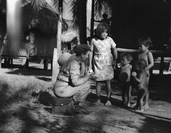 Indigenous Panamanian children talking to an American woman near the Chucunaque River in Panama. The American woman is wearing a safari hat and jumpsuit and is squatting down talking to the children. Three small boys and an older girl are standing around her, holding hands. Behind them are buildings and trees.