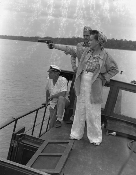 Olive Brooks, Army Division Engineer Public Relations with her .45 on the "Dolphin", Chucunaque River, Panama. She is pointing her gun out towards the river while two men are looking on. Her hair is tied up in a handkerchief and she is wearing a large button-up shirt, light-colored pants, and open toed shoes. In the background is the river and a trees along the shoreline.