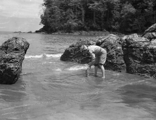 Olive Brooks, Division Engineer Public Relations, collecting dead fish after throwing dynamite into the water. She is holding a fish and has her pants rolled up to her knees. Her hair is wrapped in a handkerchief and behind her are rocks and trees. This photograph was taken on the Chucunaque River in Panama.