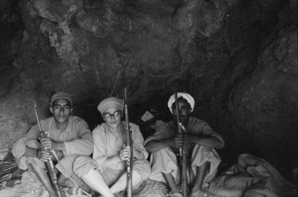 Photographer Dickey Chapelle sits with Algerian members of the National Liberation Front dressed as a member of the FLN. She is wearing a head scarf and holding a rifle while seated in a cave.