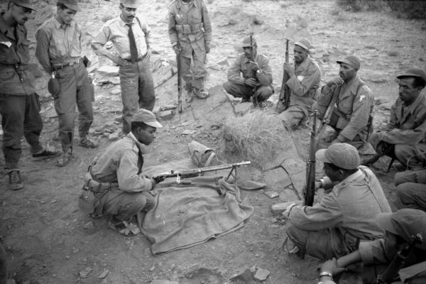 An Algerian member of the National Liberation Front showing off a gun to other FLN members. The man is sitting in the middle of a semi-circle with the gun's stand on a blanket in front of him.