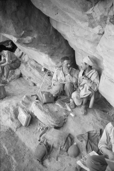 Algerian members of the National Liberation Front sitting and talking in a cave. At their feet are shoulder bags, papers, water canteens and a radio.