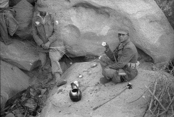 An Algerian member of the National Liberation Front drinking tea outside a cave. He is sitting on a large boulder with his gun across his lap. In front of him is a teapot and more cups. There is another man leaning against a boulder and looking down at the remains of a campfire.