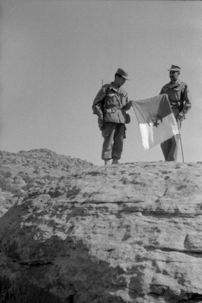 Two Algerian National Liberation Front members stand atop a boulder holding the Algerian flag between them. They are wearing fatigues and one of them is holding a cane.