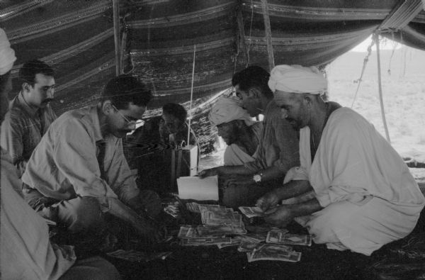 Algerian National Liberation Front members conducting a meeting in a tent. The men are sorting through papers and talking. There is a radio beside them.