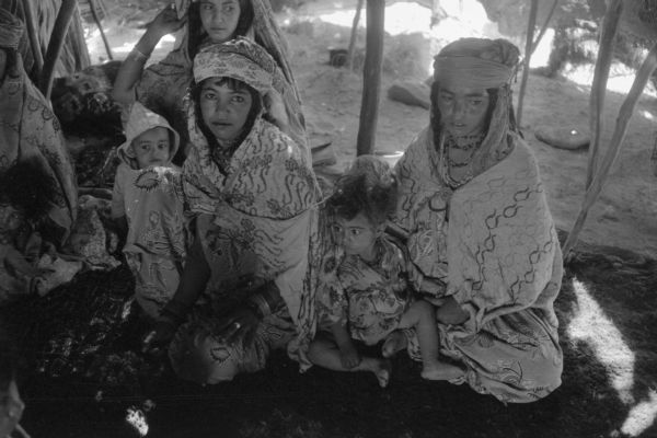 Algerian women and children gather at the Red Crescent (Red Cross equivalent) tent. They are dressed in patterned fabric and are sitting on the ground.