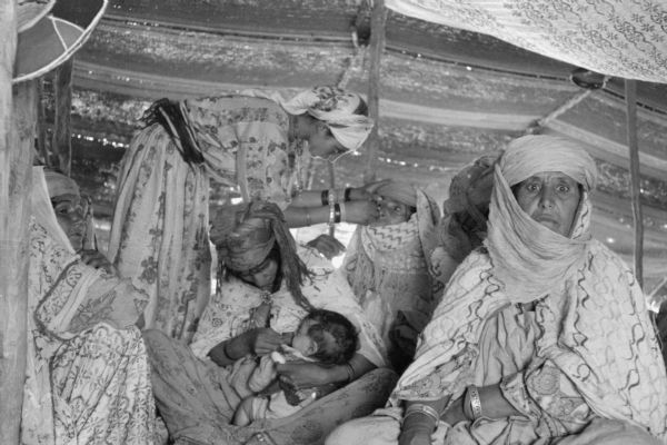 Algerian women receiving treatment in a Red Crescent (Red Cross equivalent) tent. One woman is nursing a baby. Another woman is standing while examining another woman's eye.