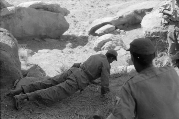 Algerian National Liberation Front member doing push-ups among boulders during training in the desert. Around him other FLN members are watching.