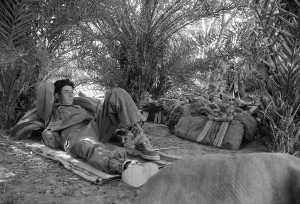 An Algerian member of the Freedom Liberation Front resting under trees. He is lying on a blanket. Near him are a pile of bags.
