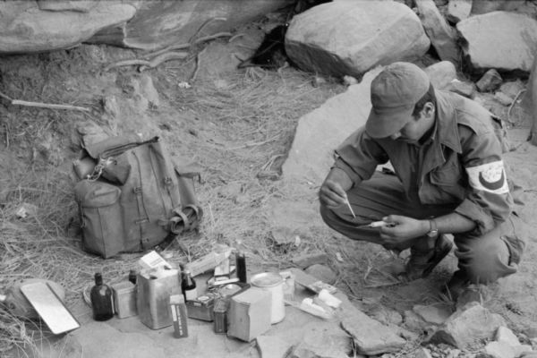 An Algerian Red Crescent (Red Cross equivalent) worker preparing a syringe while squatting on the ground near boulders. In front of him is a collection of first aid supplies. He is wearing a red crescent arm band and a cap.
