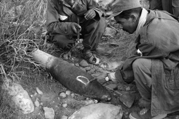 Algerian National Liberation Front members show an unexploded bomb to Dickey Chapelle, proving NATO's involvement in the war. The men are crouched around the bomb and are pointing at it.