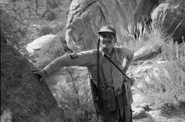An Algerian National Liberation Front member poses in the Algerian Desert. He is leaning against a boulder and squinting.