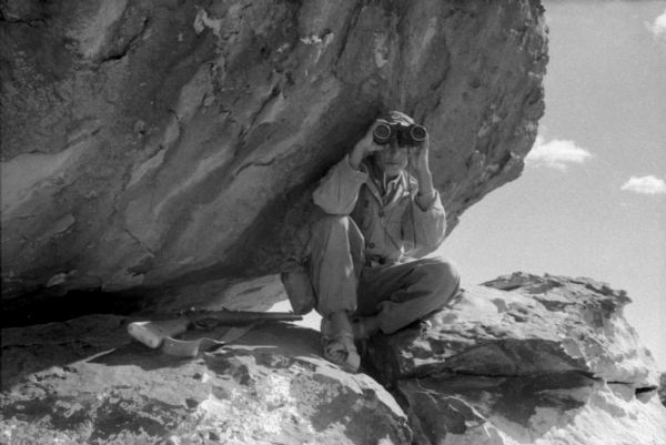An Algerian National Liberation Front member uses binoculars while on patrol. He is sitting on a boulder with his back against another boulder. Beside him is his rifle.