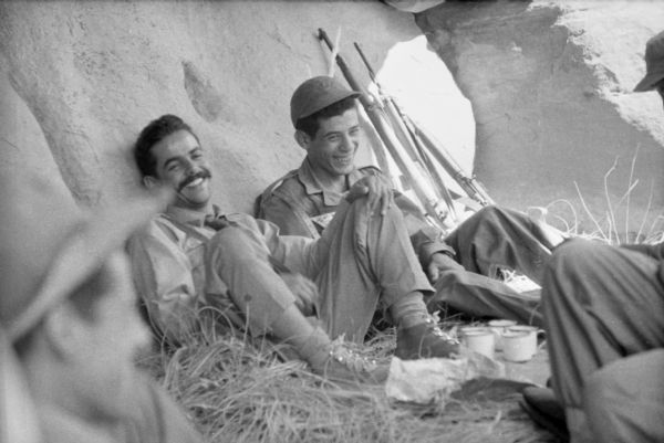 Algerian National Liberation Front members laughing while having tea. Two men are leaning against a large boulder inside a cave. There are cups at their feet and rifles beside them.