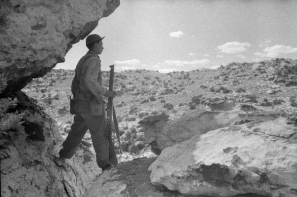 An Algerian National Liberation Front member on patrol. He is holding a rifle and straddling two boulders. Behind him is the Algerian landscape.