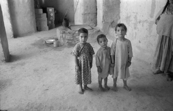 Three Algerian children standing together near their home. Two of them are holding hands. Behind them is a well, and piles of boxes in an alcove. Beside them leaning against a wall is a woman watching them.