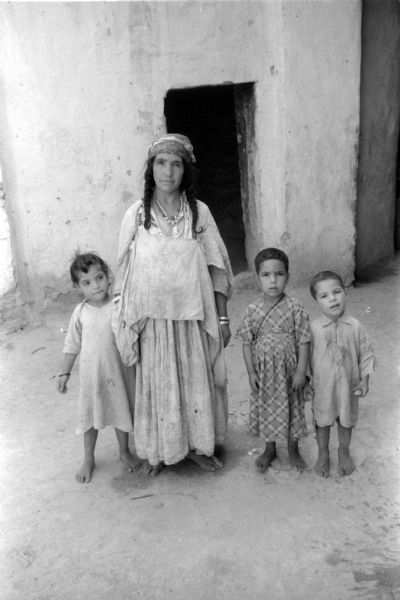 An Algerian woman with braids posing in front of a dwelling with three children. The family is barefoot, and one of the children is holding two rocks.