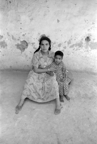 Two Algerian children sitting outside, posing for a photograph. An older girl with braids in her hair is sitting with her arm around a younger child. They are both wearing patterned dresses.