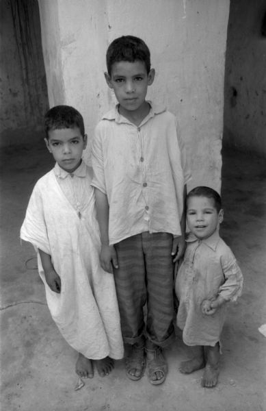 Three Algerian boys posing outdoors near a column. The tallest boy, wearing striped pants, a button up shirt, and sandals, is standing in the middle of the other two boys. The smallest boy is to the right, barefoot and wearing a long button-up shirt and holding something in his hand. The boy to the left is also barefoot and is wearing a tunic over a button-up shirt.