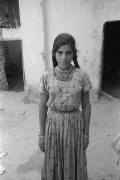 A teenage Algerian girl posing outdoors. The girl is wearing two necklaces and bracelets. She has her hair in braids and is wearing a flowered dress. She is standing in front of a dwelling with open doorways.