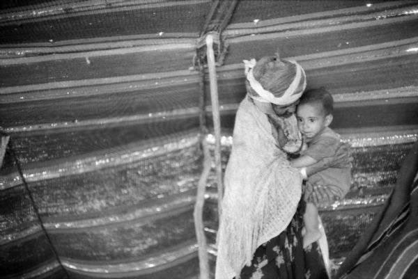 An Algerian woman is holding a baby in a tent. The woman is wearing a shawl, a turban, and a patterned dress. The tent is made of striped cloth.