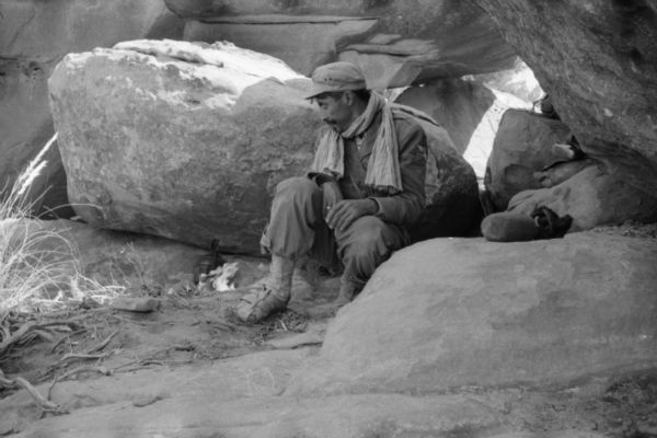 An Algerian National Liberation Front member watches a tea kettle over a fire. He is sitting in between two boulders and has a scarf around his neck.