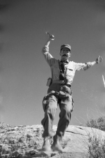 An Algerian member of the National Liberation Front poses as 'attacking' Dickey Chapelle from a boulder in the desert. He is jumping off a boulder with a large knife raised above his head.