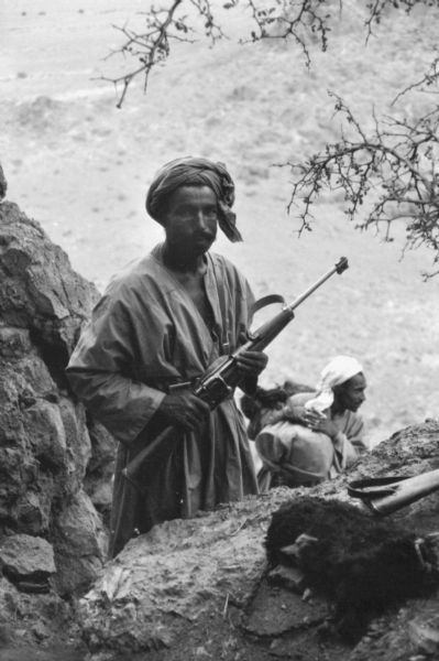 An Algerian National Liberation Front member on patrol, looking for enemy French forces. He is holding a rifle and wearing a turban and a tunic. Behind him is a man carrying a large bundle, and below him is a valley.