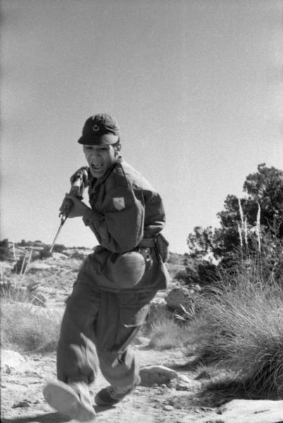 A National Liberation Front member wearing fatigues is pretending to attack Dickey Chapelle in the Algerian Desert. He is running and holding his bayonet in both hands, and there is a water canteen strapped to his belt.