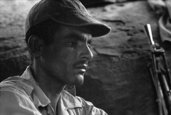 Close-up of an Algerian National Liberation Movement member resting in a cave. He is wearing a cap and a button-up shirt, and a firearm leans against a boulder in the background.