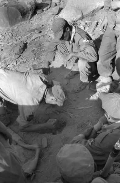 Algerian National Liberation Front members practice what it would be like to set a mine in the field. One man is digging a hole while a group of men watch him.