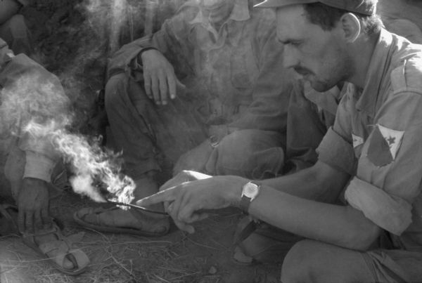 An Algerian National Liberation Front member showing other members a burning stick. He is wearing a cap and a shirt with the FLN insignia on it. He is pointing with one hand to the burning stick that he is holding with his other hand.